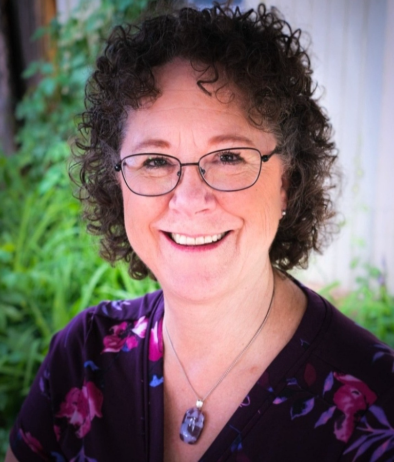 Close-up portrait of Deri, smiling warmly, wearing glasses and a purple floral top with a necklace, set against a natural outdoor background.