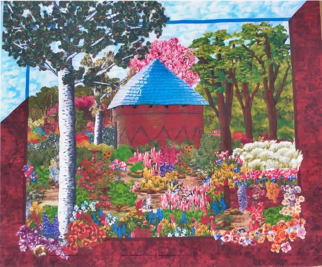 Colorful quilt art by Deri depicting a lush garden scene with a red brick building and blue roof surrounded by vibrant flowers, trees, and greenery