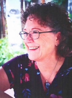 Portrait of Deri, smiling warmly and looking off-camera, wearing glasses and a floral top, with a natural outdoor background.